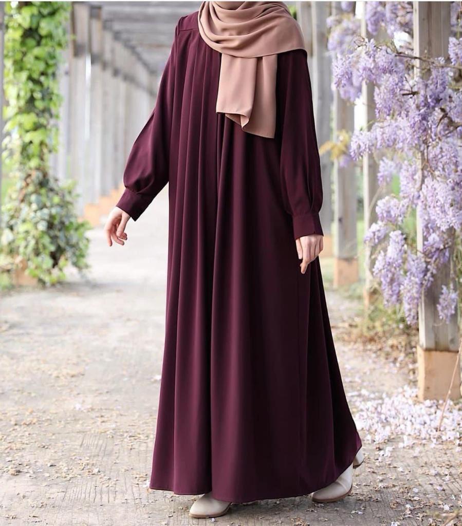 Person wearing a long burgundy dress with a beige hijab standing outdoors near flowers.