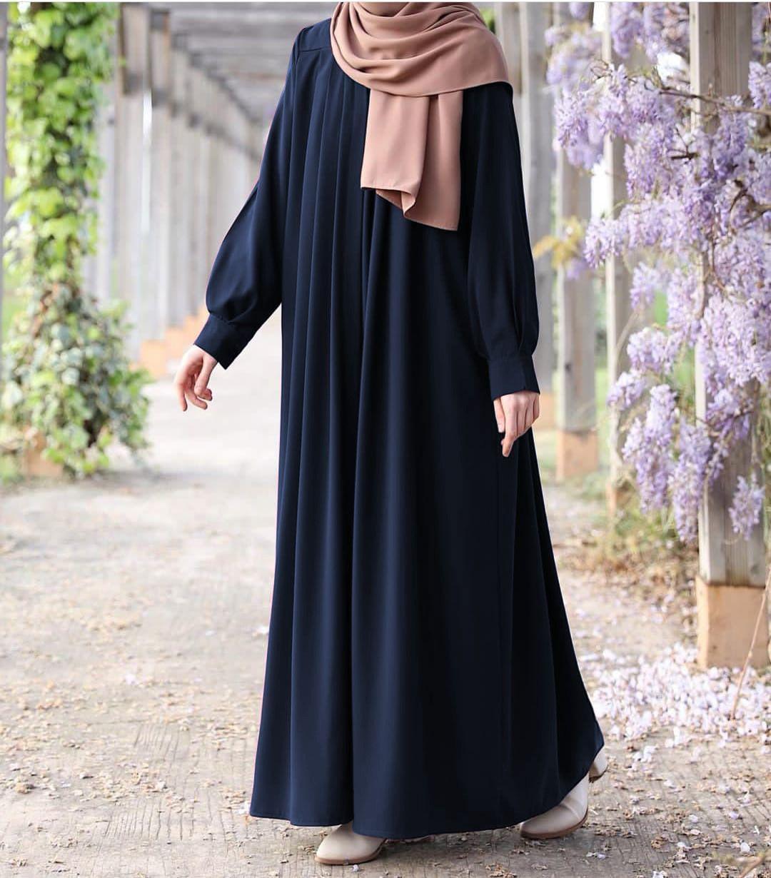 Person wearing a long dark blue dress with a beige hijab in an outdoor setting with flowers.