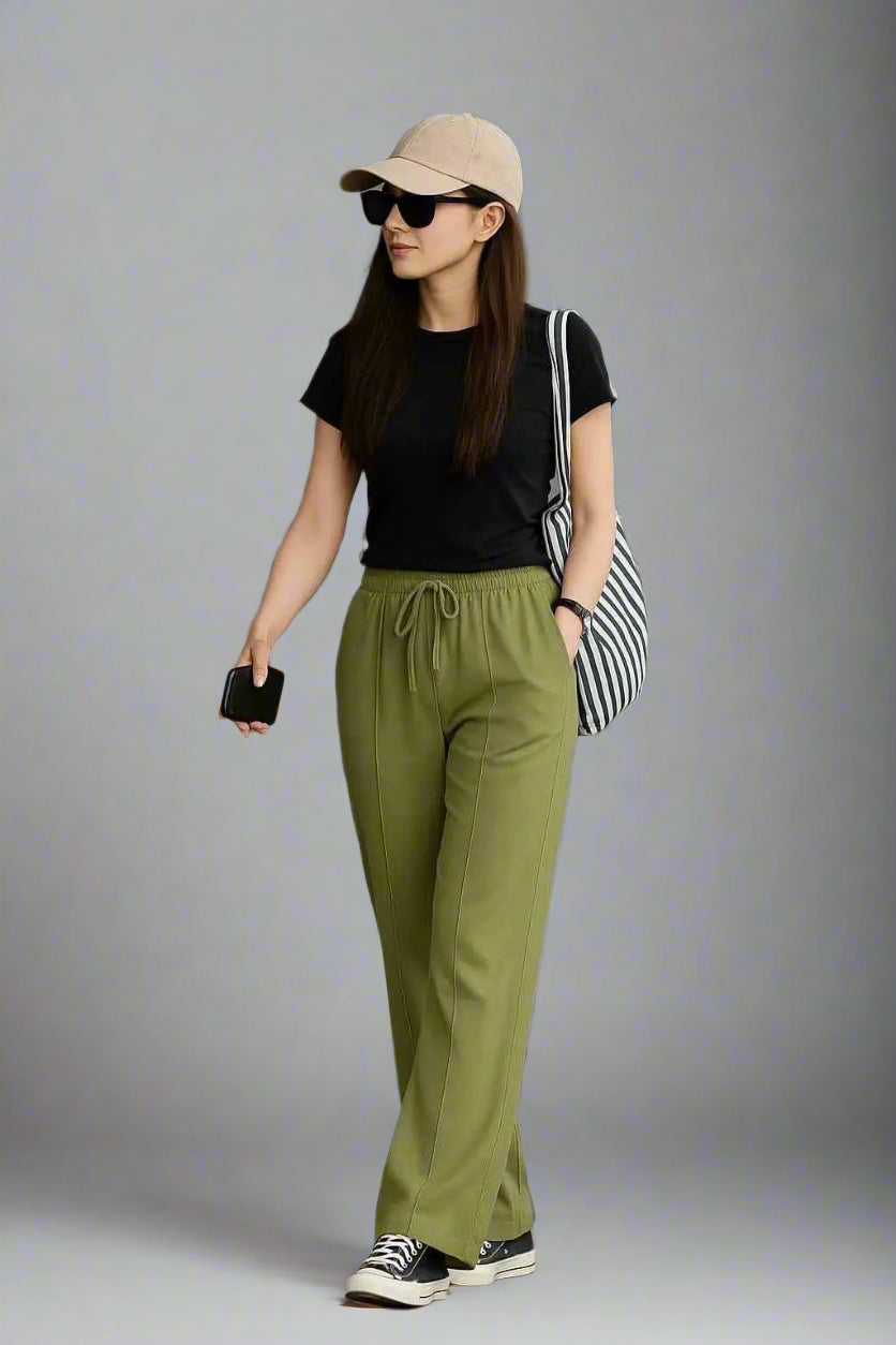 Woman wearing a black t-shirt, green pants, beige cap, and sunglasses against a gray background.